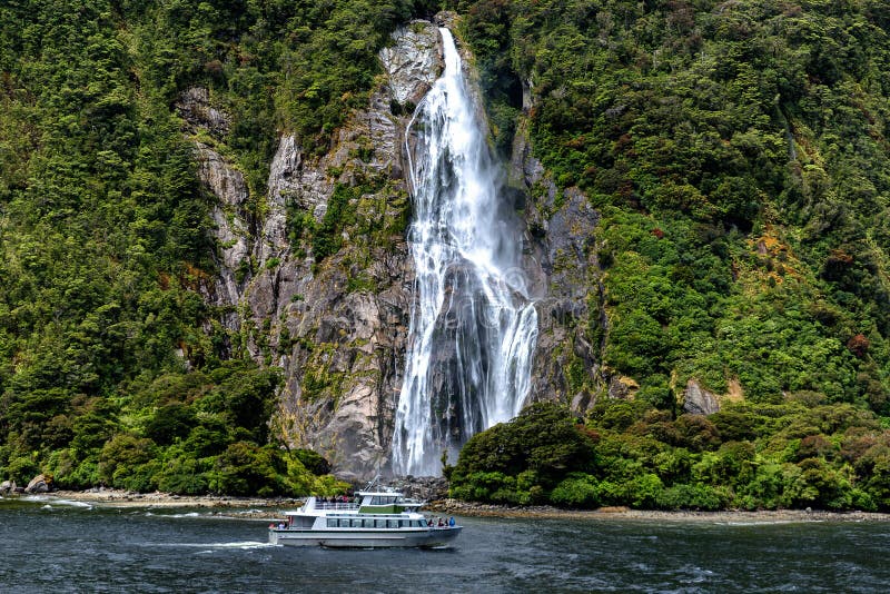Kreuzfahrtfähre und schöner hoher Wasserfall in Milford Sound, Nationalpark Fiordland lizenzfreie stockfotografie Kreuzfahrtfähre und schöner hoher Wasserfall in Milford Sound, Nationalpark Fiordland lizenzfreie stockfotografie