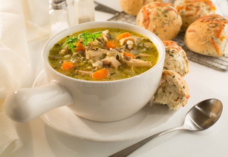 Crock of hearty chicken and wild rice soup with carrots and homemade herb and cheese rolls in white dishes on a white background. Crock of hearty chicken and wild rice soup with carrots and homemade herb and cheese rolls in white dishes on a white background