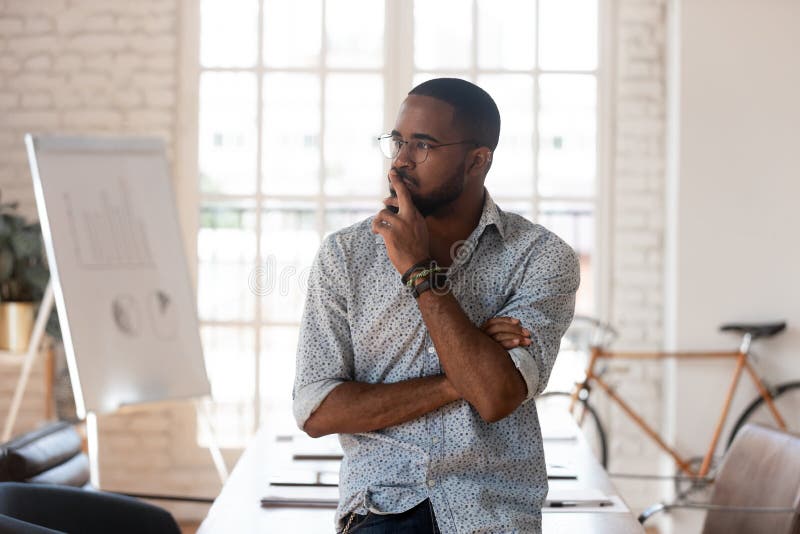 Pensive concerned african employee touch chin looking away leaned on table thinking inside of modern office boardroom, company owner boss search solution solve business issues or make decision concept. Pensive concerned african employee touch chin looking away leaned on table thinking inside of modern office boardroom, company owner boss search solution solve business issues or make decision concept