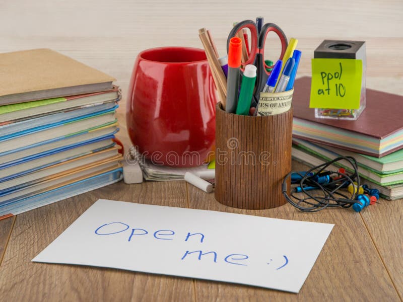 Student desktop, books, accessories, stationery, mug, in the foreground mysterious envelope with the words `open me`, front view. Student desktop, books, accessories, stationery, mug, in the foreground mysterious envelope with the words `open me`, front view