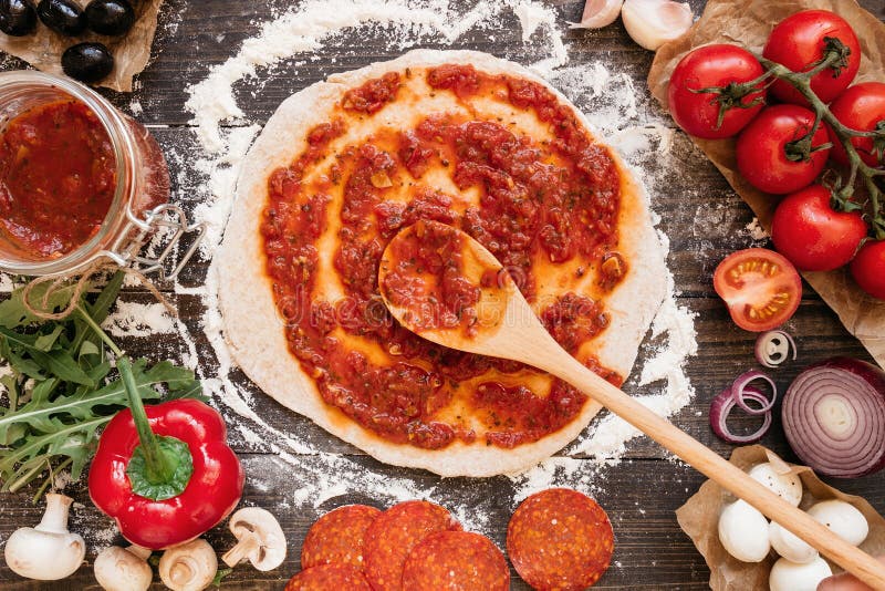 Cooking pizza. Adding fresh tomato sauce to pizza dough. Pizza ingredients on the wooden table top view. Cooking pizza. Adding fresh tomato sauce to pizza dough. Pizza ingredients on the wooden table top view.