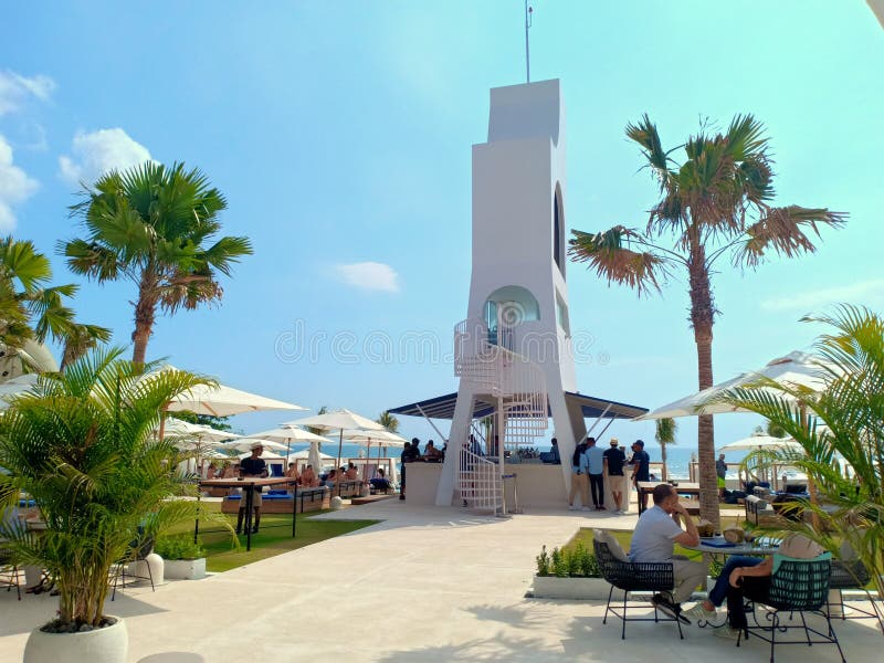 Kuta, Indonesia - 15th October 2019; Blue sky on sunny day, green palm trees, and a white tower. Tourists have a good time and relax under white umbrellas in the Cafe Del Mar Bali beach club on a sunny day. Kuta, Indonesia - 15th October 2019; Blue sky on sunny day, green palm trees, and a white tower. Tourists have a good time and relax under white umbrellas in the Cafe Del Mar Bali beach club on a sunny day