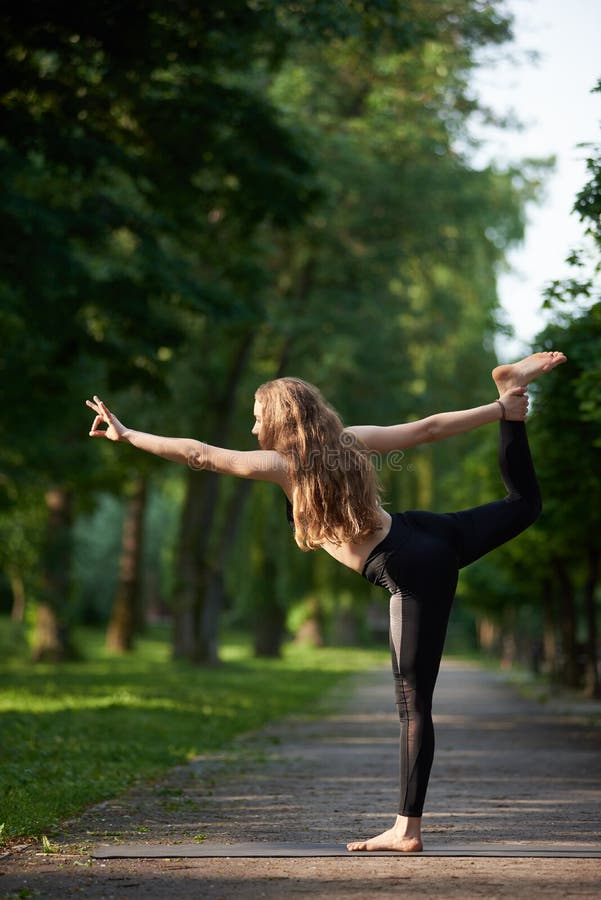 Kvinna med långt hår som står i en ställning som danskung, Asan Natarajasana mot naturparken under soliga strålar arkivbild Kvinna med långt hår som står i en ställning som danskung, Asan Natarajasana mot naturparken under soliga strålar arkivbild