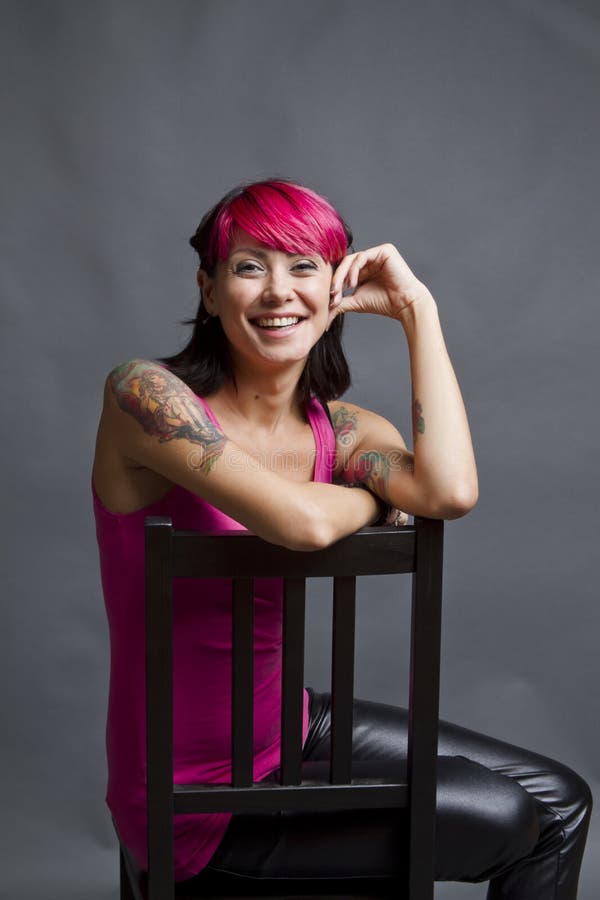 Happy, smiling woman sitting in a chair with bright pink hair, leather pants, tattoos and a big smile. Happy, smiling woman sitting in a chair with bright pink hair, leather pants, tattoos and a big smile.