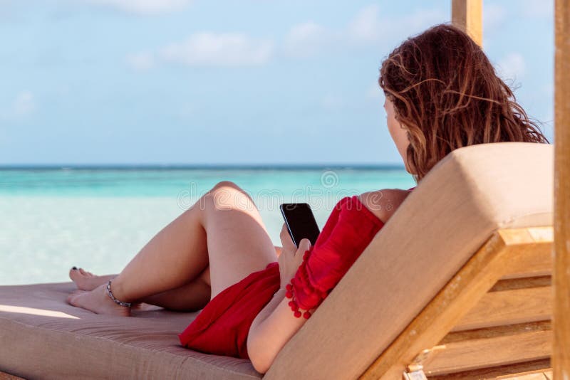 Kvinna på en sunchair i ett tropiskt läge genom att använda hennes smartphone Klart turkosvatten som bakgrund royaltyfri fotografi Kvinna på en sunchair i ett tropiskt läge genom att använda hennes smartphone Klart turkosvatten som bakgrund royaltyfri fotografi