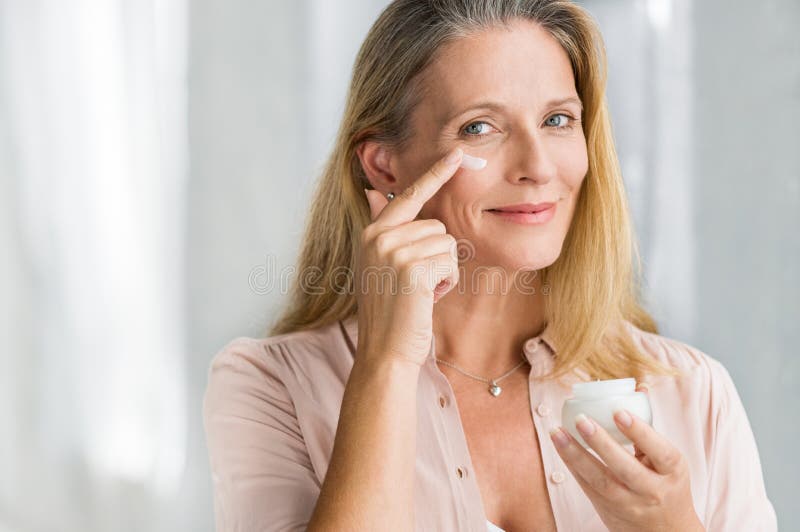 Smiling senior woman applying anti-aging lotion to remove dark circles under eyes. Happy mature woman using cosmetic cream to hide wrinkles below eyes. Lady using day moisturizer to counteract the aging of the skin. Smiling senior woman applying anti-aging lotion to remove dark circles under eyes. Happy mature woman using cosmetic cream to hide wrinkles below eyes. Lady using day moisturizer to counteract the aging of the skin.