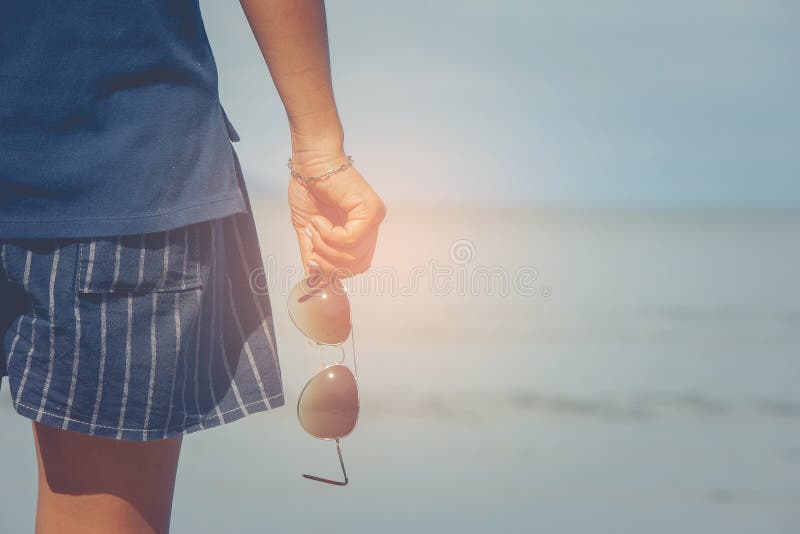 Kvinna som bär korta flåsanden och t-skjorta anseende på sandstrand- och innehavsolglasögon i hennes hand arkivbilder Kvinna som bär korta flåsanden och t-skjorta anseende på sandstrand- och innehavsolglasögon i hennes hand arkivbilder