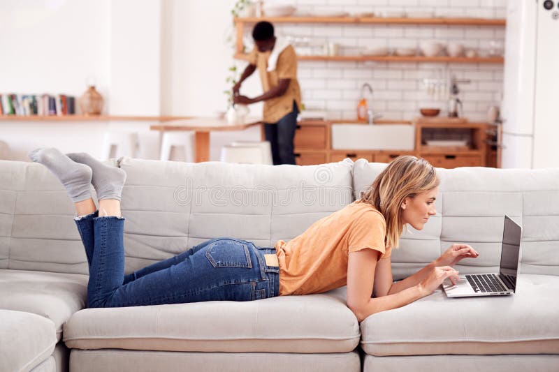 Kvinna som kopplar av att ligga på den Sofa At Home Using Laptop datoren med mannen i kök bakom arkivbilder Kvinna som kopplar av att ligga på den Sofa At Home Using Laptop datoren med mannen i kök bakom arkivbilder