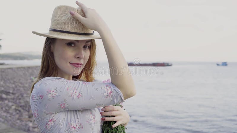 Kvinna som poserar för fotografi med hatten och blommor på havsbakgrund
