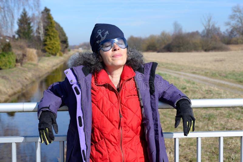 A mature woman in her early fifties wearing an outdoor jacket is standing on a bridge enjoying the Winter sun. A creek, trees and fields in the background. A mature woman in her early fifties wearing an outdoor jacket is standing on a bridge enjoying the Winter sun. A creek, trees and fields in the background.