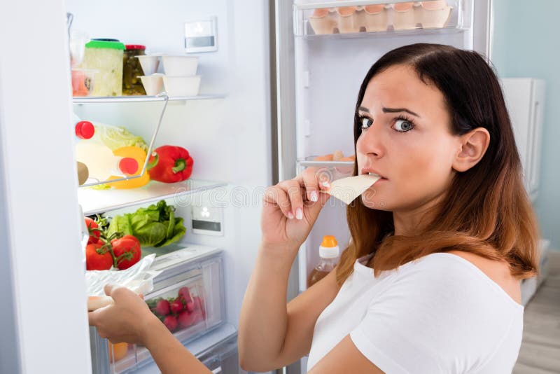 Young Woman Eating Slice Of Cheese In Front Of Open Refrigerator In Kitchen. Young Woman Eating Slice Of Cheese In Front Of Open Refrigerator In Kitchen