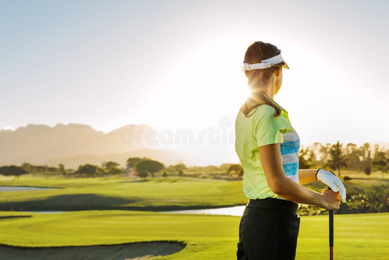 Rear view of young woman standing on golf course on a sunny day. Professional female golfer holding golf club on field and looking away. Rear view of young woman standing on golf course on a sunny day. Professional female golfer holding golf club on field and looking away.