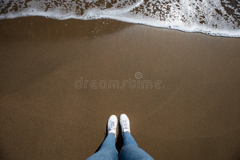 Kvinnafoten beskådar med jeans från ovannämnt på stranden med sand och vågen som kommer i ramen arkivbild Kvinnafoten beskådar med jeans från ovannämnt på stranden med sand och vågen som kommer i ramen arkivbild