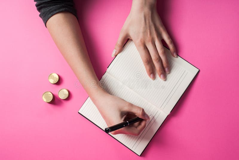 Kvinnahandhandstil med en penna i en anteckningsbok och myntet på rosa bakgrund arkivfoto Kvinnahandhandstil med en penna i en anteckningsbok och myntet på rosa bakgrund arkivfoto