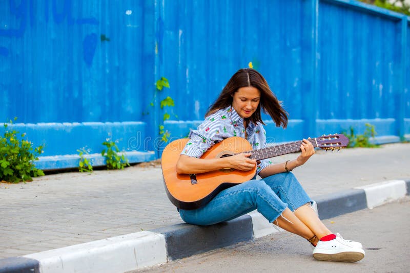 Kvinnan i jeans sitter på en vägtrottoarkant och spelar gitarren royaltyfri foto Kvinnan i jeans sitter på en vägtrottoarkant och spelar gitarren royaltyfri foto