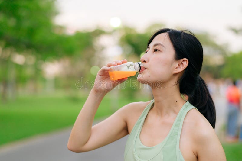 Kvinnlig joggare. lämplig ung asiatisk kvinna med grönt sportkläder som dricker ekologisk apelsinjuice efter att ha gått och njuti royaltyfri bild