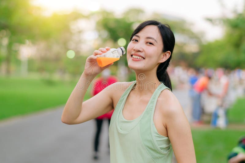 Kvinnlig joggare. lämplig ung asiatisk kvinna med grönt sportkläder som dricker ekologisk apelsinjuice efter att ha gått och njuti royaltyfria bilder