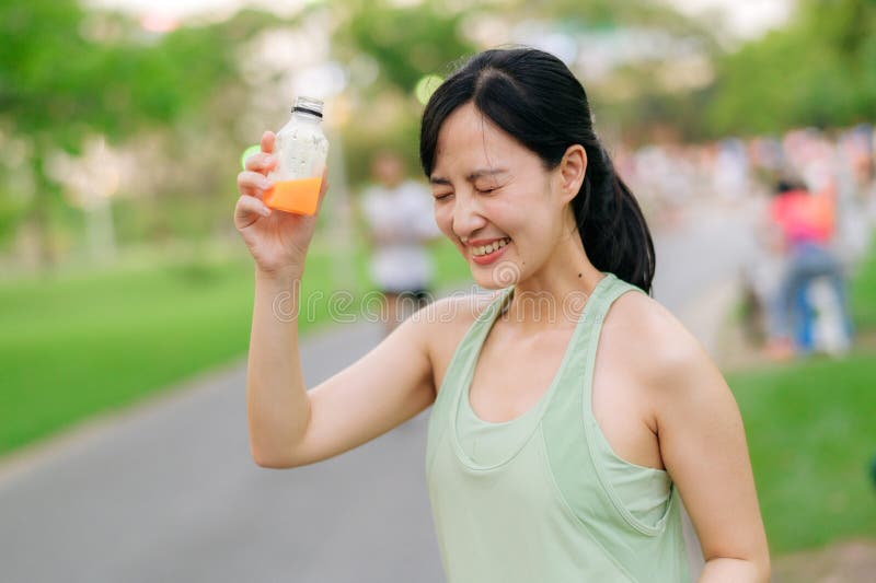 Kvinnlig joggare. lämplig ung asiatisk kvinna med grönt sportkläder som dricker ekologisk apelsinjuice efter att ha gått och njuti royaltyfri bild