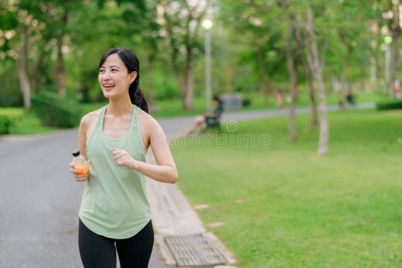 Kvinnlig joggare. lämplig ung asiatisk kvinna med grönt sportkläder som dricker ekologisk apelsinjuice efter att ha gått och njuti fotografering för bildbyråer