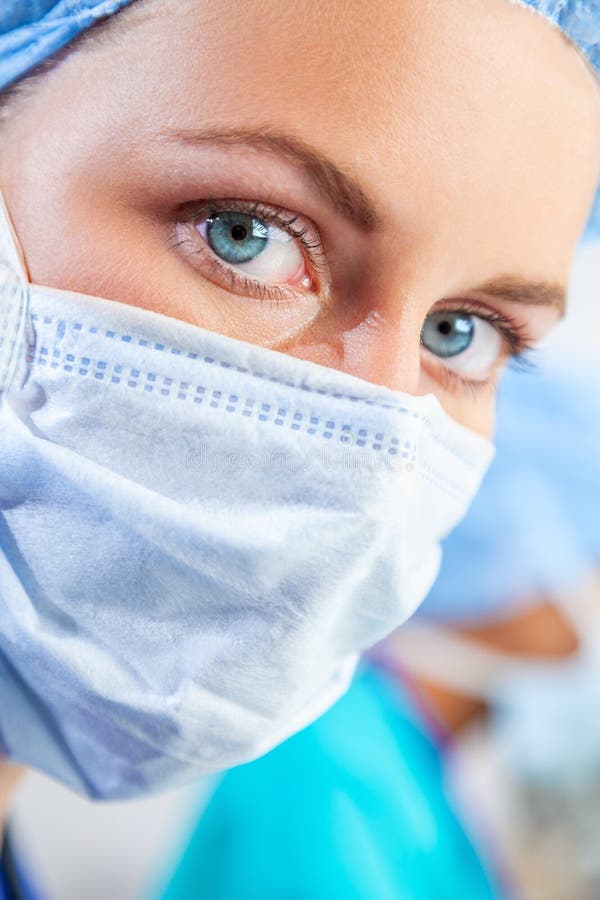Female woman doctor surgeon wearing scrubs and surgical mask in a hospital or operating theater with her colleague behind her. Female woman doctor surgeon wearing scrubs and surgical mask in a hospital or operating theater with her colleague behind her