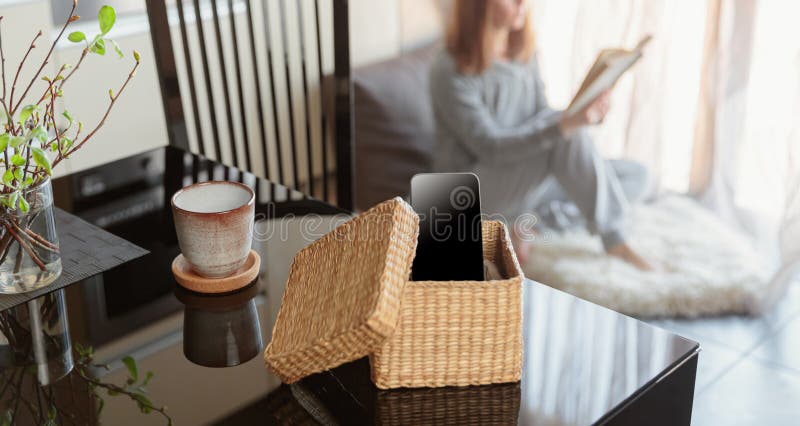 Woman sitting near window and reading book. Focus on box with smartphone. Digital detox and leisure concept. Woman sitting near window and reading book. Focus on box with smartphone. Digital detox and leisure concept