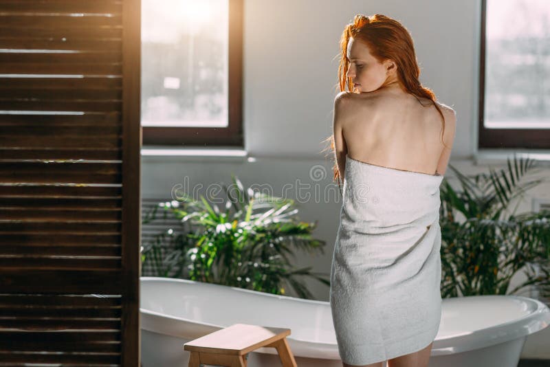 Kvinnlig modell som står mellan fönster och badkar med hjälp av handduk för att dölja sin nakenhet