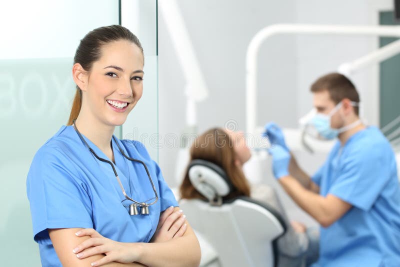 Dentist female with crossed arms wearing blue coat posing and looking at you at consultation with a doctor working with a patient in the background. Dentist female with crossed arms wearing blue coat posing and looking at you at consultation with a doctor working with a patient in the background