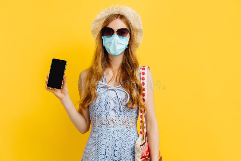 Kvinnor i sommarhatt och medicinsk mask som visar en tom smartphone-skärm och pekar på den medan man står mot gul bakgrund royaltyfria foton