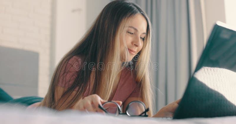 Kvinnor som skriver på den bärbara datorn i sängen i sovrummet. tar bort glasögon och placerar det på tangentbordet