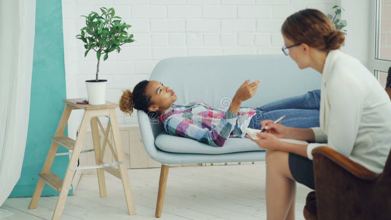 L'adolescent d'afro-américain parle au psychologue se trouvant sur le divan et souriant tandis que le thérapeute écoute et prend banque de vidéos
