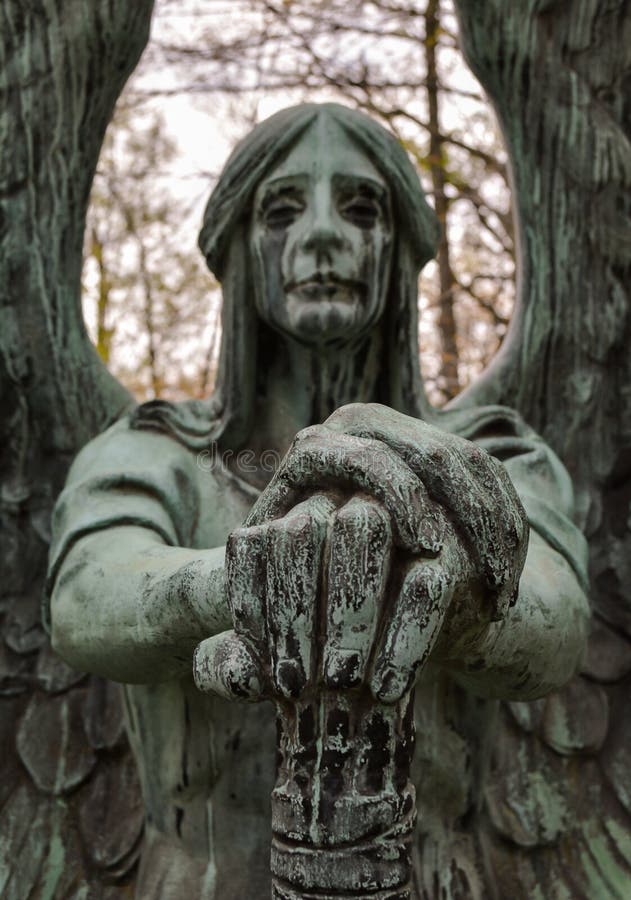 An eerie closeup view of an angel statue at a grave site who appears to be weeping black tears down its face and neck due to the effects of aging and weathering of the bronze its made of. Its wings are stretched out behind it, and the image has a 3D affect with its hands focused in front, resting on an upside down torch, extinguishing life. An eerie closeup view of an angel statue at a grave site who appears to be weeping black tears down its face and neck due to the effects of aging and weathering of the bronze its made of. Its wings are stretched out behind it, and the image has a 3D affect with its hands focused in front, resting on an upside down torch, extinguishing life.