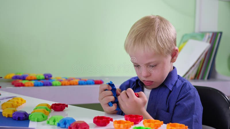 L'enfant concerné avec la trisomie 21 désassemble et joue avec le constructeur coloré assis à une table à la maison étroite banque de vidéos