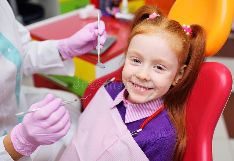 L'enfant est une séance de sourire de petite fille rousse dans une chaise dentaire image stock L'enfant est une séance de sourire de petite fille rousse dans une chaise dentaire image stock