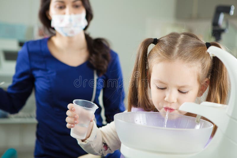 Child rinses out mouth and spit in special sink, holds plastic cup with water in hand and doctor in work uniform and sterile mask sits behind in modern medical office. Child rinses out mouth and spit in special sink, holds plastic cup with water in hand and doctor in work uniform and sterile mask sits behind in modern medical office.