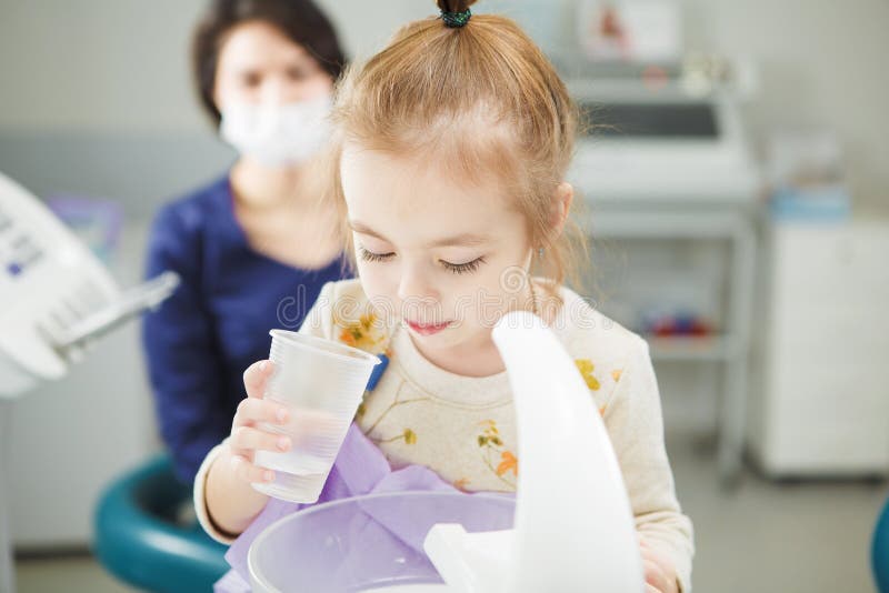 Child rinses out mouth and spit in special sink, holds plastic cup with water in hand and doctor in work uniform and sterile mask sits behind in modern medical office. Child rinses out mouth and spit in special sink, holds plastic cup with water in hand and doctor in work uniform and sterile mask sits behind in modern medical office.