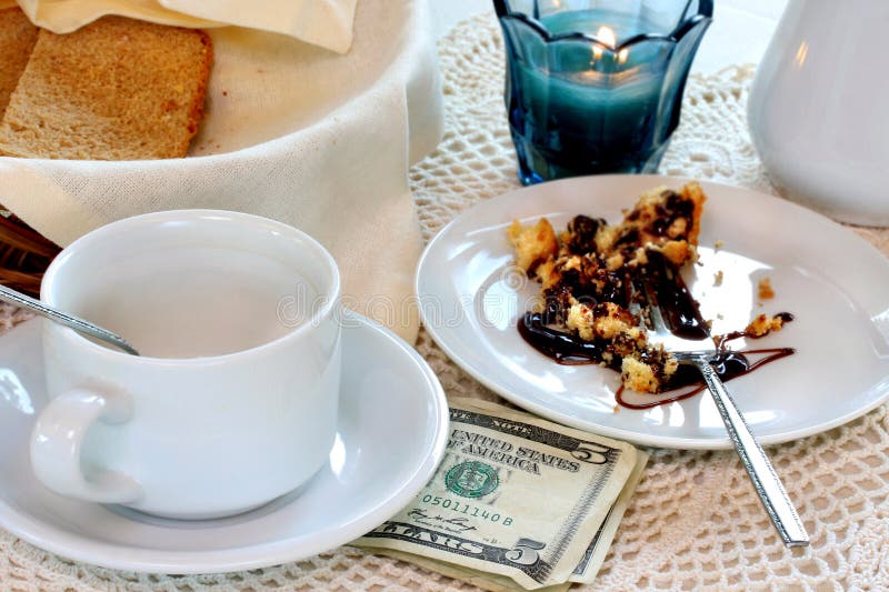 A five dollar bill with other bills sits under an empty coffee cup on a table with other items indicating a finished meal. A five dollar bill with other bills sits under an empty coffee cup on a table with other items indicating a finished meal.