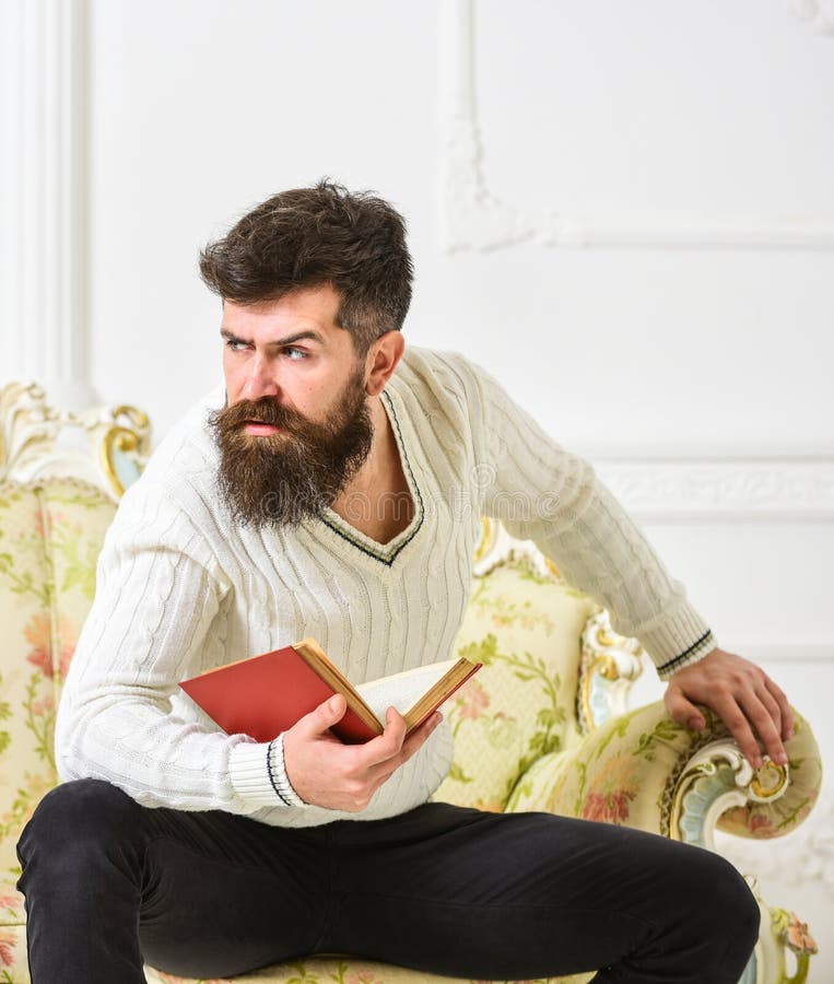 L'homme avec la barbe et la moustache s'assied sur le sofa baroque de style, tient le livre, fond blanc de mur Réflexions sur la photo stock L'homme avec la barbe et la moustache s'assied sur le sofa baroque de style, tient le livre, fond blanc de mur Réflexions sur la photo stock