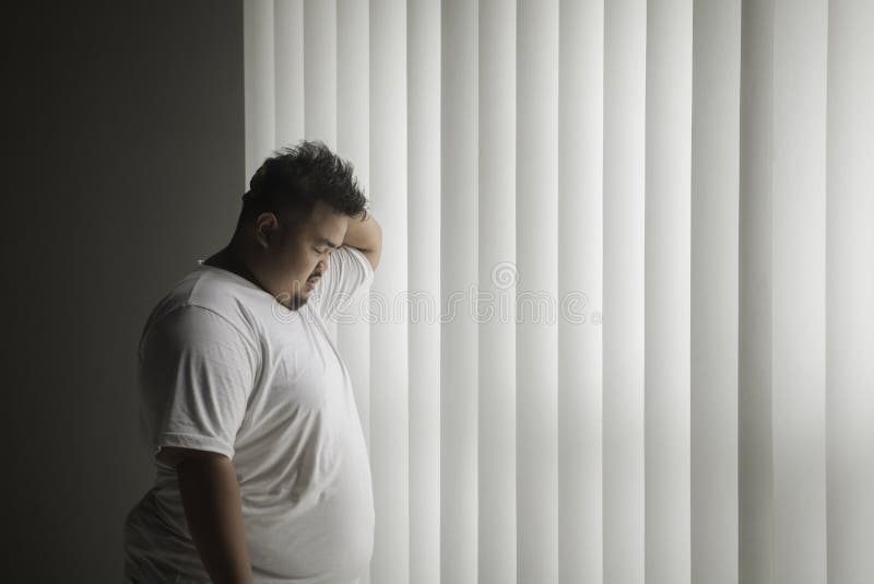 Picture of overweight man looks lonely while standing near the window in the dark room. Picture of overweight man looks lonely while standing near the window in the dark room