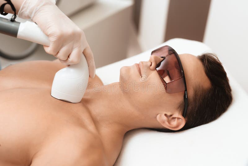 The man came to the procedure of laser hair removal. The doctor treats his neck and face with a special apparatus. The man has red glasses. The man came to the procedure of laser hair removal. The doctor treats his neck and face with a special apparatus. The man has red glasses.