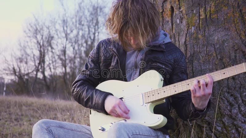 L'homme joue la guitare électrique dans un domaine près de l'arbre au coucher du soleil