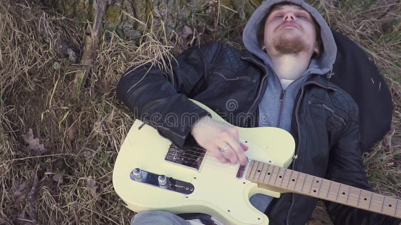 L'homme joue la guitare électrique dans un domaine près de l'arbre au coucher du soleil