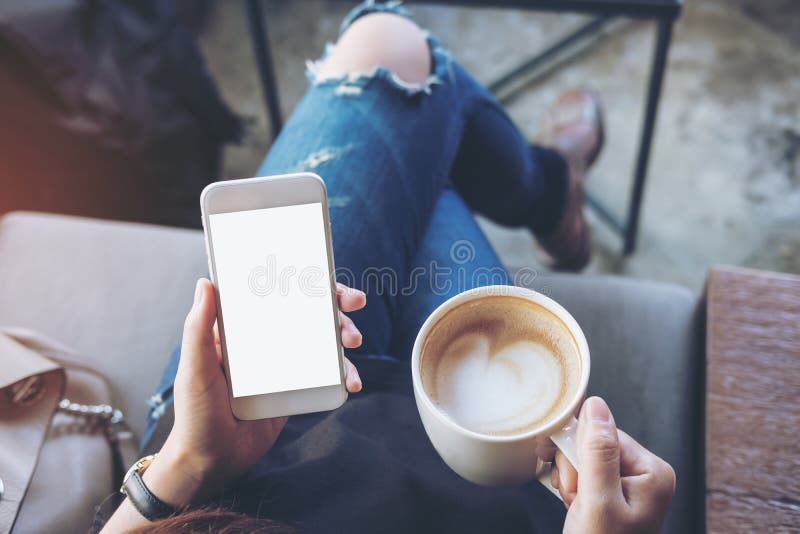 Mockup image of woman`s hands holding white mobile phone with blank screen on thigh and coffee cup in cafe. Mockup image of woman`s hands holding white mobile phone with blank screen on thigh and coffee cup in cafe