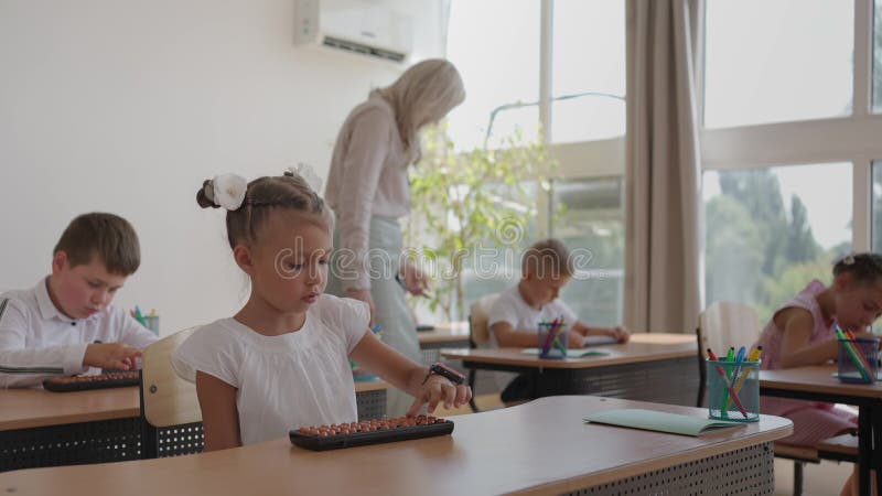 L'instituteur se promène dans la salle de classe de l'école. un groupe d'enfants est assis à leur bureau. les enfants écrivent banque de vidéos