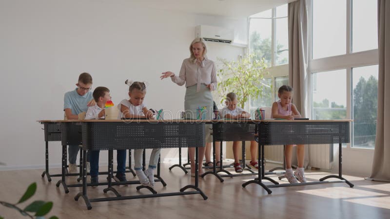 L'instituteur se promène dans la salle de classe de l'école. un groupe d'enfants est assis à leur bureau. les enfants écrivent clips vidéos