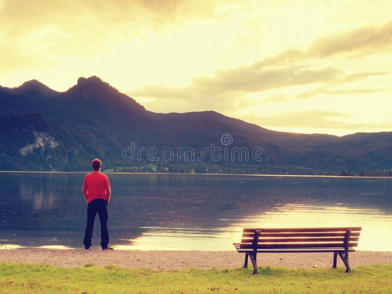 L'uomo alto in maglietta rossa al banco di legno a buio della costa del lago delle montagne si appanna immagine stock L'uomo alto in maglietta rossa al banco di legno a buio della costa del lago delle montagne si appanna immagine stock