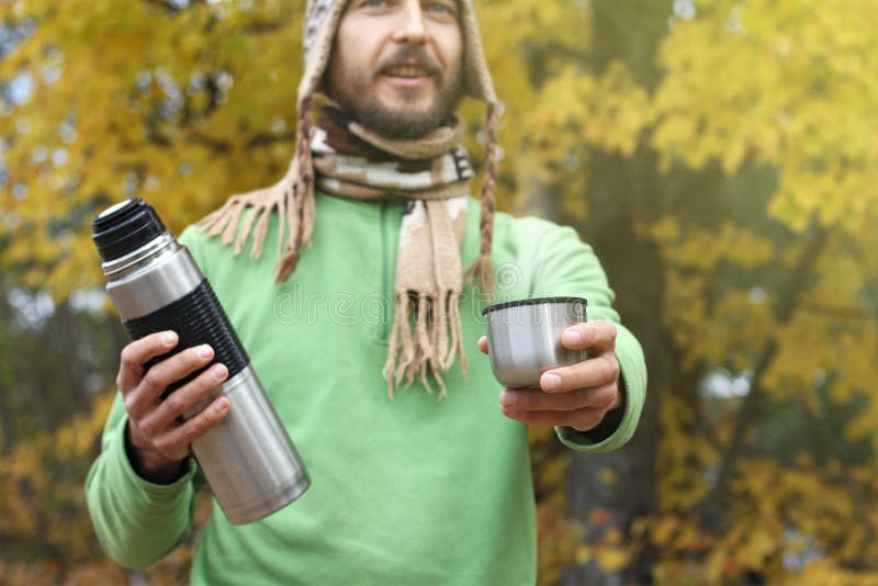 L'uomo in cappello e sciarpa tricottati, con il sorriso offre la bevanda calda - tè o caffè dal termos a qualcuno, vista frontale fotografie stock libere da diritti L'uomo in cappello e sciarpa tricottati, con il sorriso offre la bevanda calda - tè o caffè dal termos a qualcuno, vista frontale fotografie stock libere da diritti