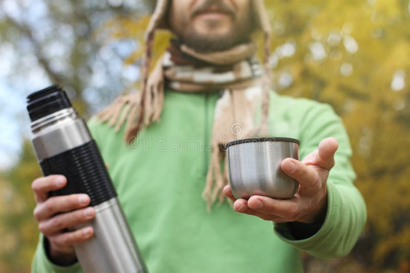 L'uomo in cappello e sciarpa tricottati, con il sorriso offre la bevanda calda - tè o caffè dal termos, vista frontale fotografie stock libere da diritti L'uomo in cappello e sciarpa tricottati, con il sorriso offre la bevanda calda - tè o caffè dal termos, vista frontale fotografie stock libere da diritti