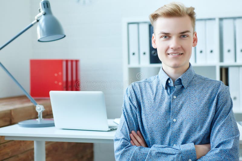 L'uomo caucasico sorridente di affari dei giovani sta esaminando la macchina fotografica, sedentesi davanti al suo posto di lavor fotografia stock libera da diritti L'uomo caucasico sorridente di affari dei giovani sta esaminando la macchina fotografica, sedentesi davanti al suo posto di lavor fotografia stock libera da diritti