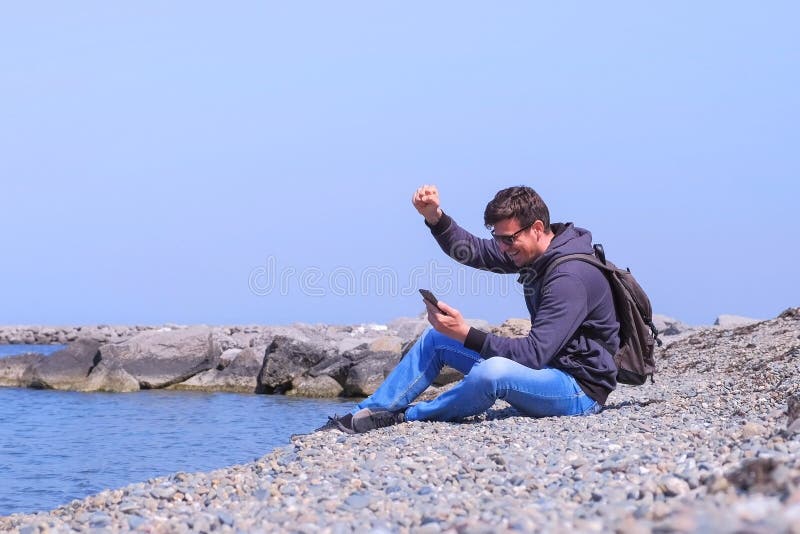 L'uomo che il viaggiatore è felice circa ottenere le grandi notizie sullo smartphone si siede alla spiaggia del mare immagini stock