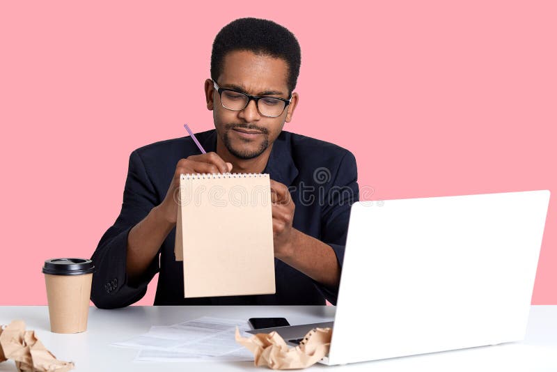 L'uomo d'affari nero premuroso prova a scrivere la poesia in taccuino per la sua amica mentre lavora al computer portatile Afroam immagini stock libere da diritti L'uomo d'affari nero premuroso prova a scrivere la poesia in taccuino per la sua amica mentre lavora al computer portatile Afroam immagini stock libere da diritti